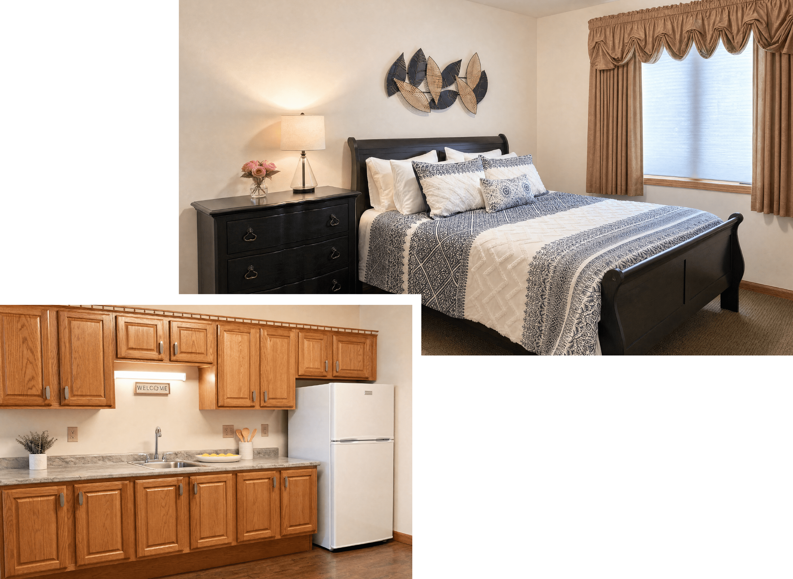 A cozy bedroom with a large bed covered in blue and white patterned bedding, decorated with multiple pillows, and a black wooden headboard. Next to the bed is a black nightstand with a lamp and a small vase of pink flowers. The room has a window with brown curtains and a decorative wall art piece above the bed. The kitchen area features wooden cabinets, a white refrigerator, a sink, and a 
