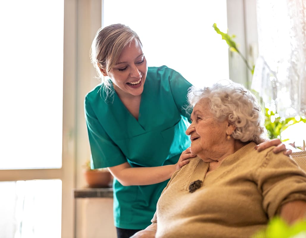 A caregiver, wearing teal scrubs, smiling and leaning towards an elderly woman with curly white hair, who is seated and smiling back. They are indoors with sunlight coming through a window, and there are green plants in the background.