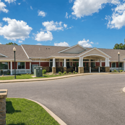 A single-story rehabilitation center building with a brick and white siding exterior, a gabled roof, and a covered entrance supported by columns. The building is set against a bright blue sky with scattered clouds, and there are lamp posts, a sidewalk, and a small grassy area in front.