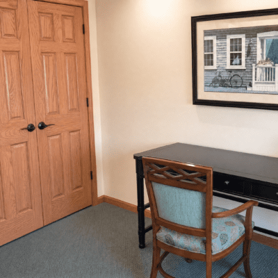 A corner of a room with a wooden door on the left, a black desk with a decorative backrest chair in front, and a framed picture of a house and bicycle hanging on the white wall.