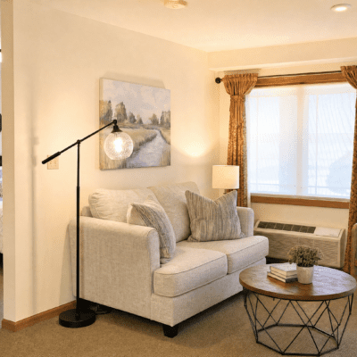A cozy living room with a white sofa adorned with several beige and gray pillows. A round wooden coffee table with a metal frame holds a potted plant and a book. To the right, a large window with white curtains and gold drapes lets in natural light. A floor lamp with a spherical glass shade stands next to the sofa, and a landscape painting hangs on the wall behind it.