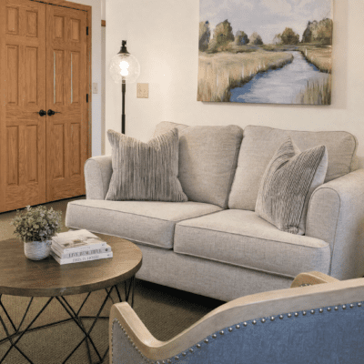 A cozy living room with a light gray sofa adorned with three matching pillows, a round wooden coffee table with a plant and books, a blue armchair with nailhead trim, a wooden door, a floor lamp, and a landscape painting of a river and trees hanging on the wall.
