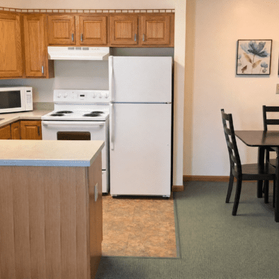 A kitchen with wooden cabinets, a white stove, a white refrigerator, and a microwave on the counter. To the right, there is a dining area with a table and four black chairs, and a framed floral artwork on the wall.