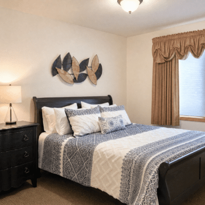 A neatly made bed with a dark wooden frame, covered with a blue and white patterned quilt and several decorative pillows. To the left of the bed is a black nightstand with a lamp. Above the bed hangs a modern, abstract metal wall art piece. There is a window with beige curtains and a valance on the right side of the room.