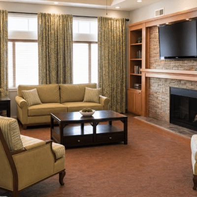 A cozy living room with a beige sofa and matching armchair, a dark wooden coffee table with a decorative bowl, large windows with patterned curtains, a brick fireplace, and a wall-mounted flat-screen TV above the fireplace.