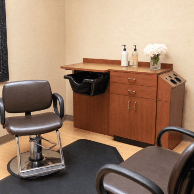A therapy or consultation room with two brown chairs, a wooden cabinet with drawers, a small sink with soap and sanitizer bottles, a flower vase, and a black trash bin.