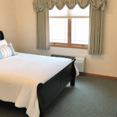 A bedroom with a black bed frame and white bedding, positioned next to a window with light green curtains and valance. The window lets in natural light, and there is a radiator below it. The room has beige walls and a gray carpeted floor.