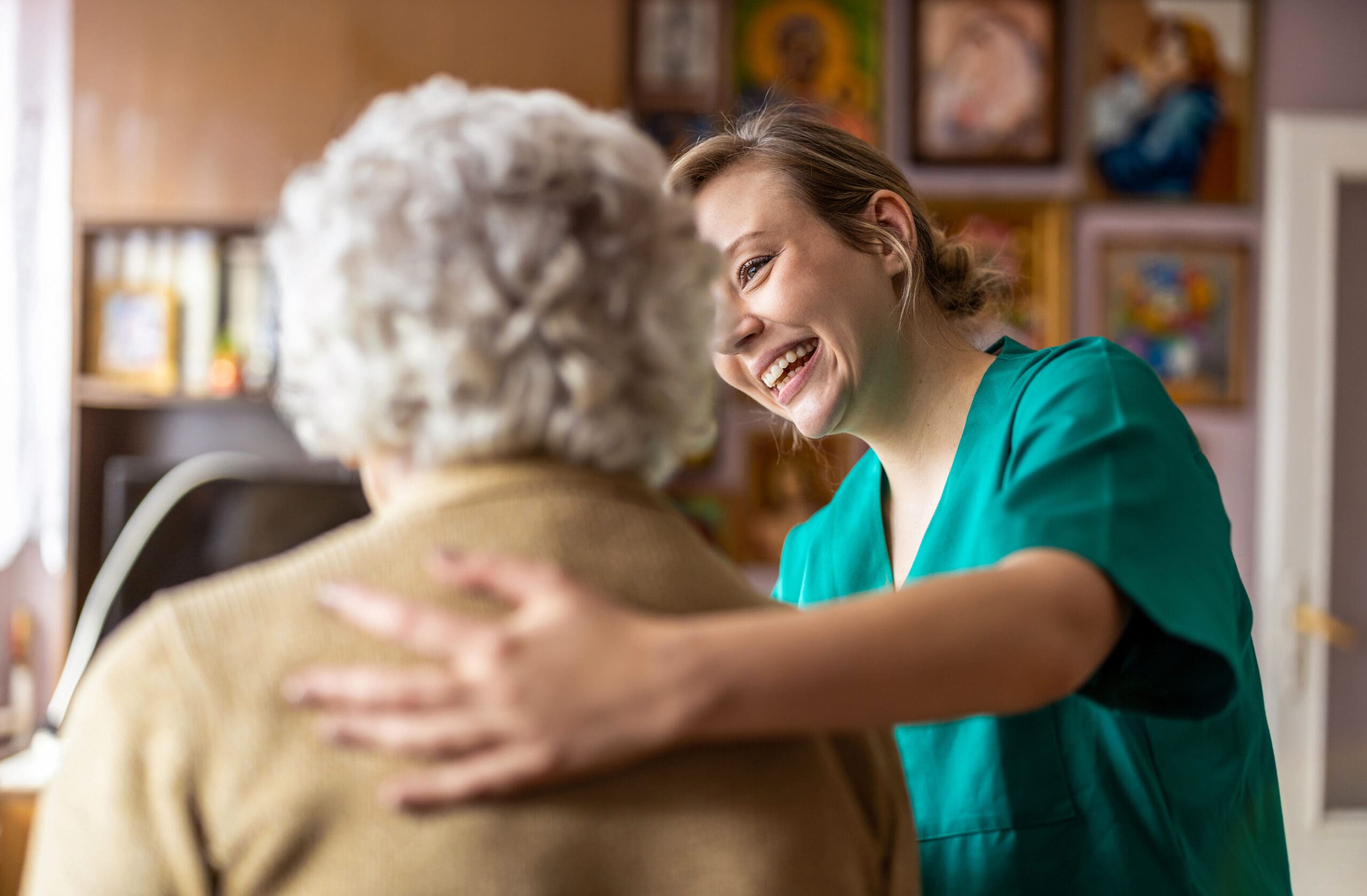 A young female healthcare worker with a bright smile embracing an elderly woman with white hair in a cozy room decorated with framed artwork.