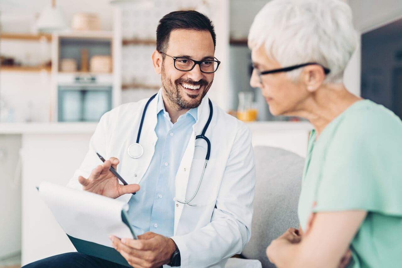 A male doctor with dark hair, glasses, and a stethoscope around his neck, smiling and talking to an elderly female patient with white hair and glasses, in a bright, modern rehabilitation or nursing center.