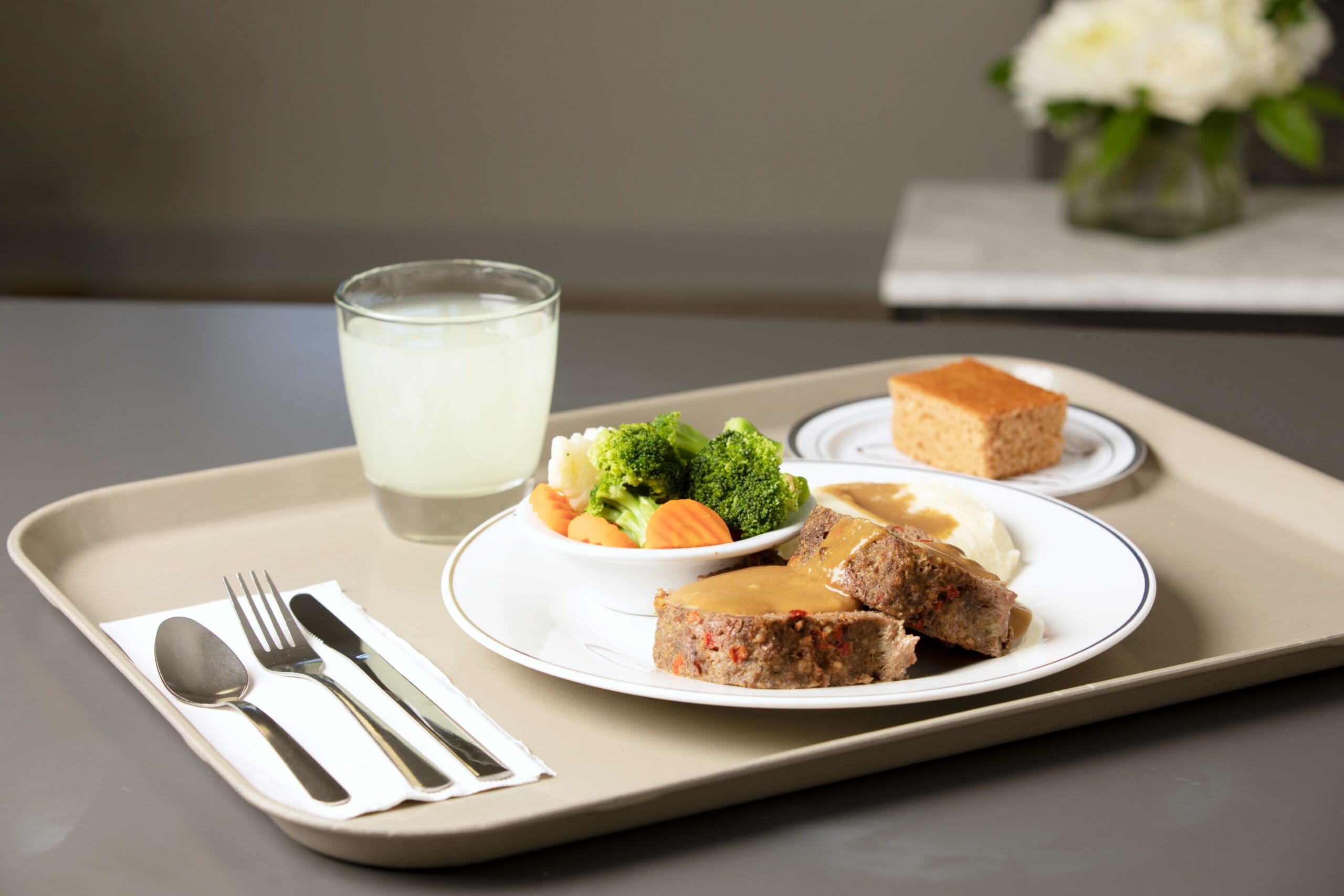 A tray with a meal including a plate of meatloaf with gravy, mashed potatoes, and a side of broccoli and carrots. There is a small bowl of broccoli and carrots, a glass of water, a piece of cake on a separate plate, and a set of utensils on a napkin.