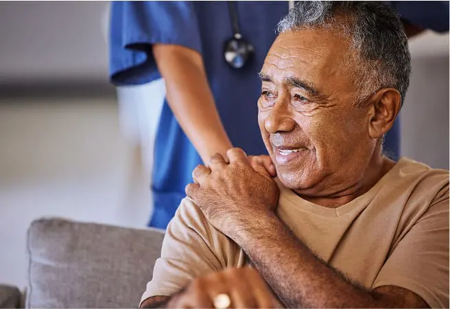 An elderly man smiling and resting his hand on his shoulder, with a healthcare worker in blue scrubs standing behind him in a medical or rehabilitation setting.