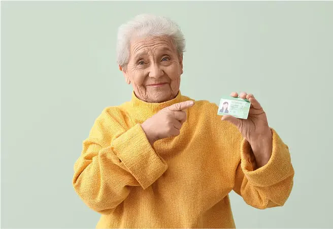 An elderly woman with white hair, wearing a yellow sweater, smiling and pointing at a small card she is holding in her right hand against a light green background.