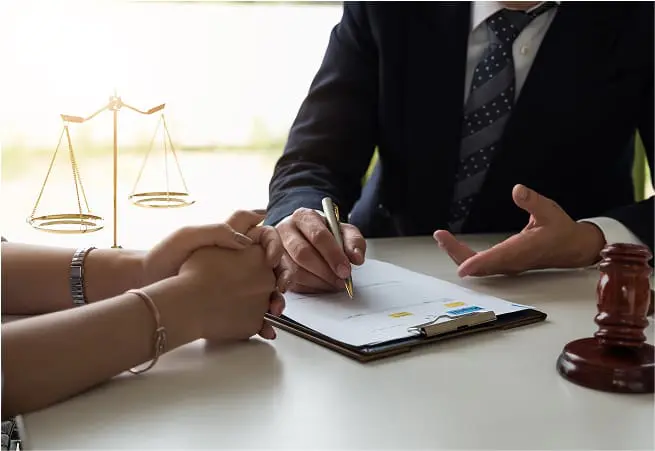 A person in a suit and tie is sitting at a desk, signing documents. Another person, whose hands are visible, is holding the signer’s hands in a gesture of reassurance. There is a gavel on the desk and a set of scales of justice in the background.