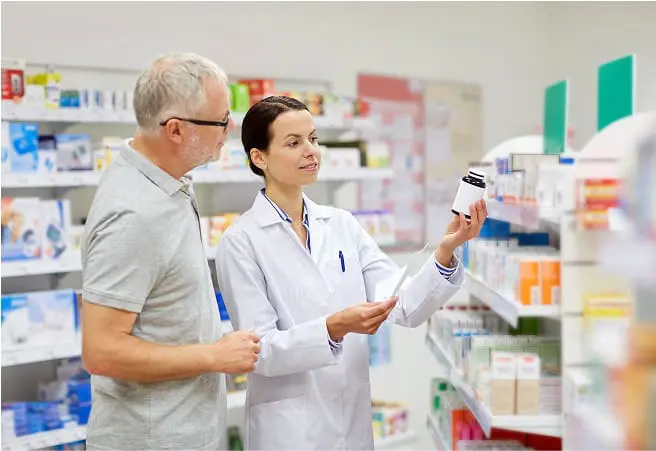 A pharmacist showing a bottle of medication to a male customer in a pharmacy aisle filled with various medicines and health products.