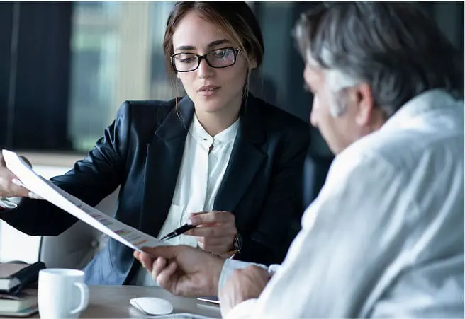 A young woman in professional attire and glasses is sitting at a desk, reviewing documents with an older man in a white shirt. They are engaged in a serious discussion in a modern office setting.