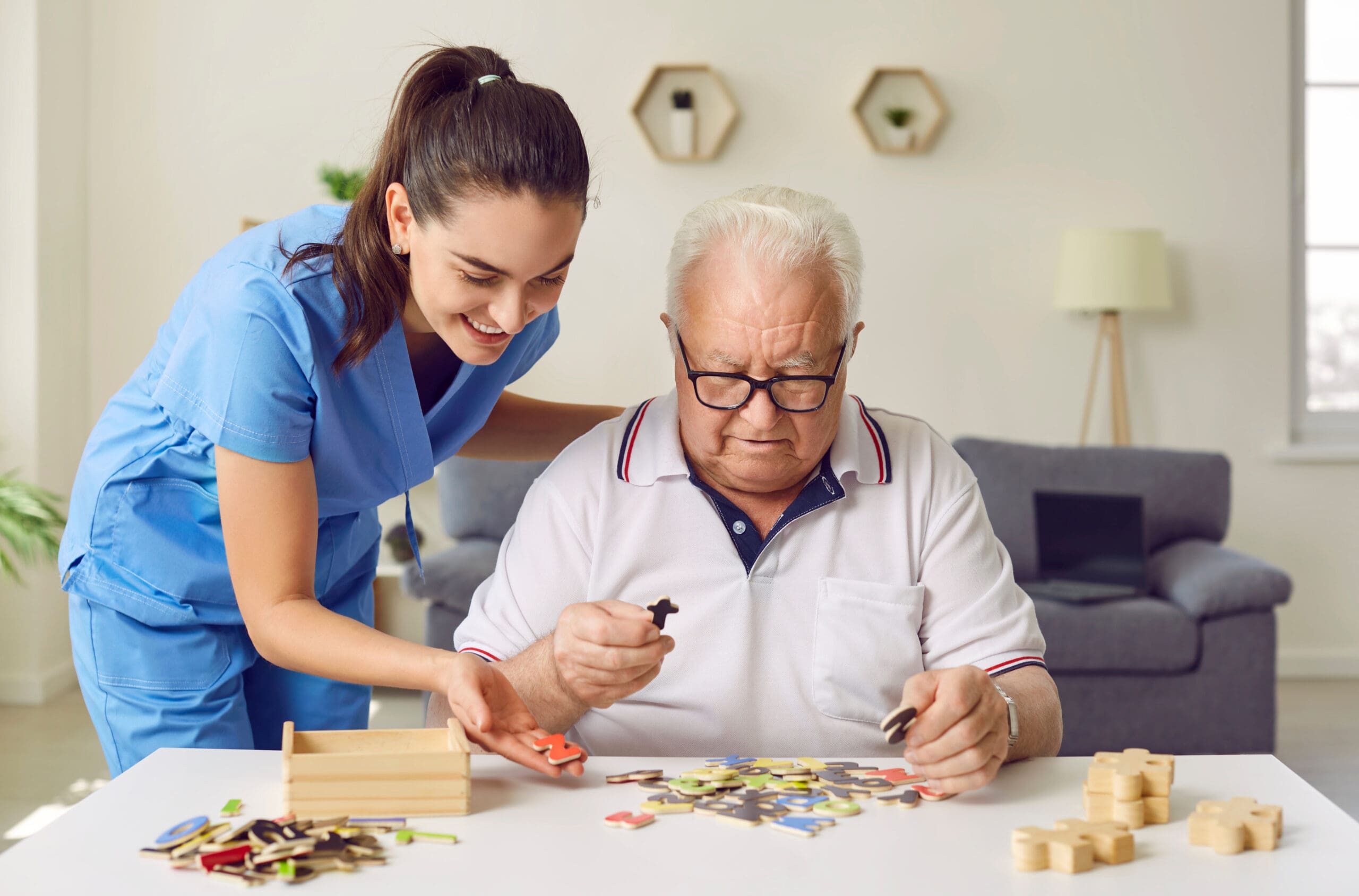A young female caregiver in blue scrubs helps an elderly man with glasses play a puzzle game at a table, smiling and engaging in a recreational activity together.